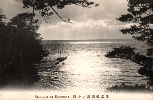 This black-and-white card shows a coastal view framed by pine trees, with boats on the water. On the card is printed “NISHIURA AT ENOSHIMA.” and “江之嶋西浦ノ夕照”. The Japanese 江之嶋西浦ノ夕照 translates as “Evening glow at Nishiura, Enoshima” or “Sunset at Nishiura, Enoshima.”