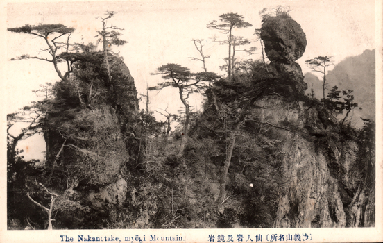 This black-and-white card shows steep rocky mountain formations with pine trees growing among the cliffs. On the card is printed “The Nakanotake, myōgi Mountain.” and “（妙義山名所）仙人岩及鏡岩”. The Japanese translates as:
妙義山名所 — “Famous sights of Mount Myōgi”
仙人岩及鏡岩 — “Sennin Rock and Kagami Rock”, literally “Hermit Rock and Mirror Rock.”