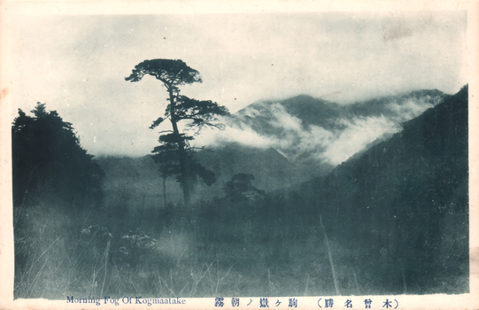 This black-and-white card shows a misty mountain landscape with a tall tree in the foreground. On the card is printed “Morning Fog Of Kogmaatake” and “木曾名勝　駒ヶ嶽ノ朝霧”. The Japanese translates in English as “Morning mist of Mount Komagatake, a famous scenic place in Kiso.”