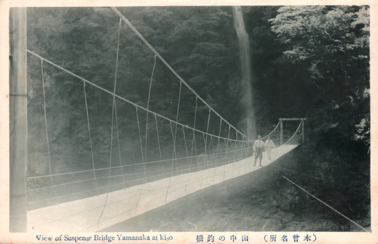 This black-and-white card shows a suspension bridge crossing a wooded ravine, with two people standing on the bridge. On the card is printed “View of Suspense Bridge Yamanaka at kiso”, “山中の釣橋” and “（木曾名所）”. The Japanese translates as:
山中の釣橋 — “Suspension bridge at Yamanaka”
木曾名所 — “Famous sights of Kiso”