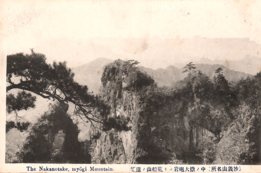 This black-and-white card shows steep rocky mountain formations with pine trees. On the card is printed “The Nakanotake, myōgi Mountain.” and “（妙義山名所）中ノ嶽大砲岩ヨリ荒船山ノ遠望”. The Japanese translates as:
妙義山名所 — “Famous sights of Mount Myōgi”
中ノ嶽大砲岩ヨリ荒船山ノ遠望 — “Distant view of Mount Arafune from Cannon Rock at Nakanotake.”