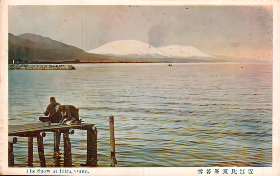 11731.200 This coloured card shows a snow-covered mountain range across a body of water, with two figures on a wooden platform in the foreground. On the card is printed “The Snow at Hira, Oumi.” and “近江比良峯暮雪”. The Japanese 近江比良峯暮雪 translates as “Evening snow on Mount Hira, Ōmi.”