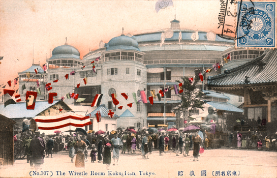 This coloured card shows a crowded street outside a large arena building decorated with flags. On the card is printed “(No.107) The Wrestle Room Kokugikan, Tokyo.”, “國技館” and “（東京名所）”. The Japanese translates as:
國技館 — “Kokugikan” / “National Sports Hall”
東京名所 — “Famous places of Tokyo” / “Tokyo sights”