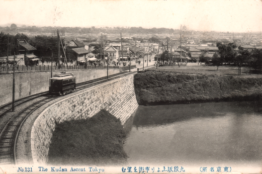 This black-and-white card shows a tram travelling beside a moat with a city streetscape in the distance. On the card is printed “No.131 The Kudan Ascent Tokyo”, “九段坂より市街を望む” and “（東京名所）”. The Japanese translates as:
九段坂より市街を望む — “View of the city from Kudan Slope”
東京名所 — “Famous places of Tokyo”