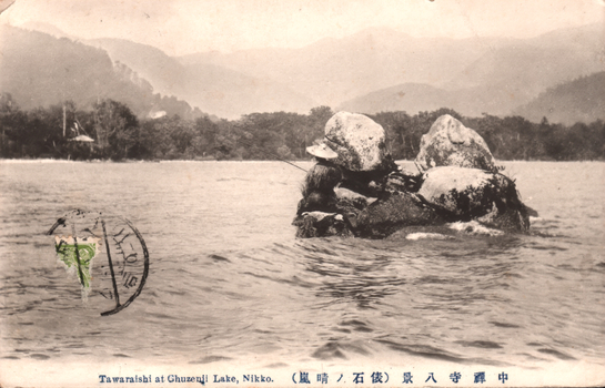 This black-and-white card shows a person beside large rocks in a lake, with wooded hills and mountains in the distance. On the card is printed “Tawaraishi at Chuzenji Lake, Nikko.”, “（嵐晴ノ俵石）” and “中禅寺八景”. The Japanese translates as:
嵐晴ノ俵石 — “Tawara Stone after the storm clears”
中禅寺八景 — “Eight Views of Chuzenji”