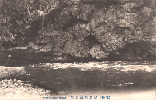This black-and-white card shows a river flowing below a steep rocky cliff. On the card is printed “KUMAKAWA HIGO.” and “球磨川鎧岩（肥後）”. The Japanese translates as:
球磨川 — “Kuma River”
鎧岩 — “Armour Rock” / “Yoroi Rock”
肥後 — “Higo”
Together, the Japanese caption means approximately: “Armour Rock on the Kuma River, Higo.”