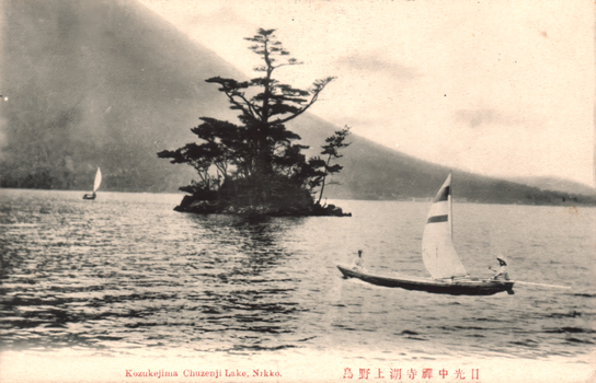 This black-and-white card shows small sailboats on a lake beside a tree-covered islet, with hills behind. On the card is printed “Kozukejima Chuzenji Lake, Nikko.” and “日光中禅寺湖上野島”. The Japanese translates as:
日光 — “Nikko”
中禅寺湖 — “Lake Chuzenji”
上野島 — “Kozukejima” / “Kōzuke Island”
Together, the Japanese caption means: “Kozukejima on Lake Chuzenji, Nikko.”