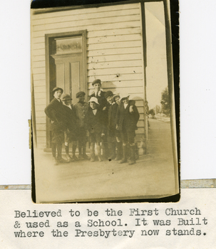 Eight boys in various shorts, walking socks,jackets and caps standing as a group in front of a small weather board building.
