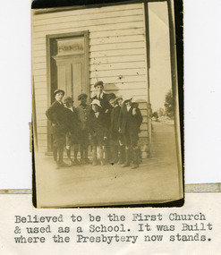 Eight boys in various shorts, walking socks,jackets and caps standing as a group in front of a small weather board building.
