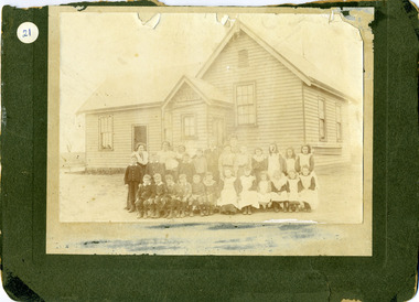 Group of students and teachers standing outside School a weatherboard building.In three rows girls on the right, boys on the left. 