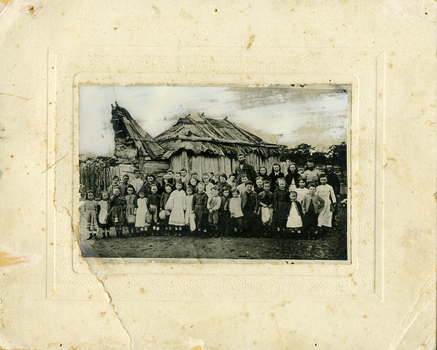 43 students and their teacher Mr Nicholson standing in front of bark hut school. Children are of various ages.