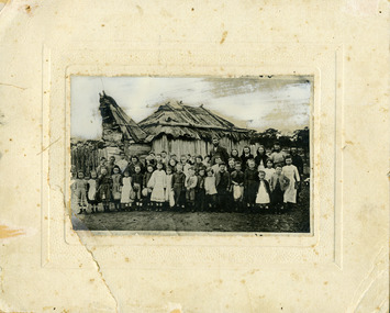 43 students and their teacher Mr Nicholson standing in front of bark hut school. Children are of various ages.