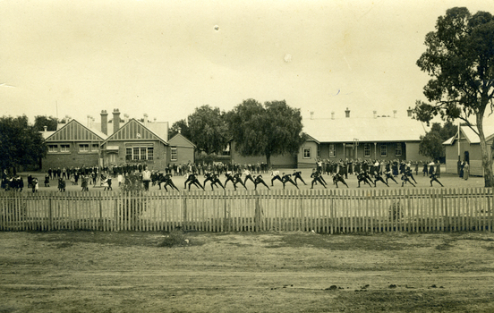 A large group of students exercising in front of the school while other students watch. Picket fence in the foreground.
