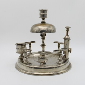 Silver stand to hold 8 pipes and pipe cleaner, A bell on top. Match striker plate and cigar cutter. Candle holder on top. Tarnished.