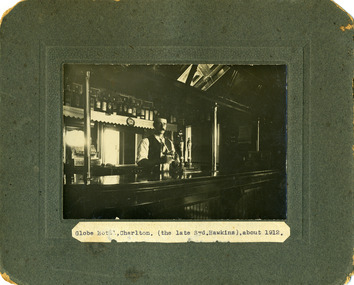 Man standing behind the bar cleaning a glass. Polished wooden bar. Bartender has a mostach, vest and tie. Rows of spriit bottles behind.
