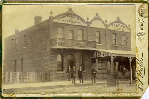 Looking from High Street Charlton at the Commonwealth Bank and Procter and Son butchers. Four men standing in front of the Bank and 2 more in front of Procter's, one in a butcher apron. Both buildings are brick with large signage.