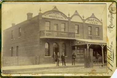 Looking from High Street Charlton at the Commonwealth Bank and Procter and Son butchers. Four men standing in front of the Bank and 2 more in front of Procter's, one in a butcher apron. Both buildings are brick with large signage.