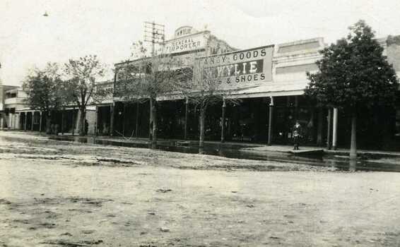 Flooded main street. Main shop front in Willey's Store. Wide varanda with two children standing underneath with flood water lapping at the pavement.