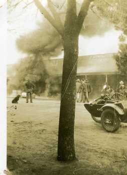 Petrol bowser fire. Onlookers include a dog and a man, three men standing together, and three other men on the rh side of the photo. A motorbike with a side car.