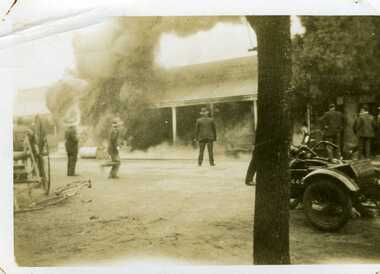 c. 1930 Garage fire. Thick smoke bellowing from building. Cart and motorbikes on street. Six men watch from the street.