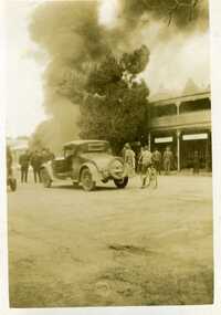Petrol bowser fire c.1930, thick smoke bellowing from building. Crowd of people watching from the street. Car and boy on bike parked in street.