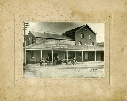 Six men with motorcycles standing in front of the Charlton Motor P/L service station. Various signs on the buliding with a large post veranda.