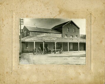 Six men with motorcycles standing in front of the Charlton Motor P/L service station. Various signs on the buliding with a large post veranda.