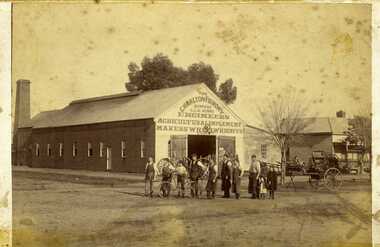 Ten people standing outside. Long timber building, Machinery, and horse drawn vehicles on display. 