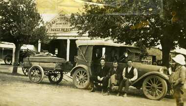  A Car and trailer are parked in front. Three men are sitting on the car running board and two men are inside the car. A boy is standing at the right of the photo.