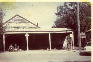 An older couple and small dog sit in front of Charlton Foundry building. The building has a large varanda and wooden construction. Sign reads "Charlton Foundry Company, Engineers"