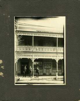 Three men standing under the varanda of the State Bank, they are all dressed in suits. The building is two storey brick with wrought iron decoration on the varanda.