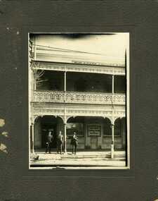Three men standing under the varanda of the State Bank, they are all dressed in suits. The building is two storey brick with wrought iron decoration on the varanda.