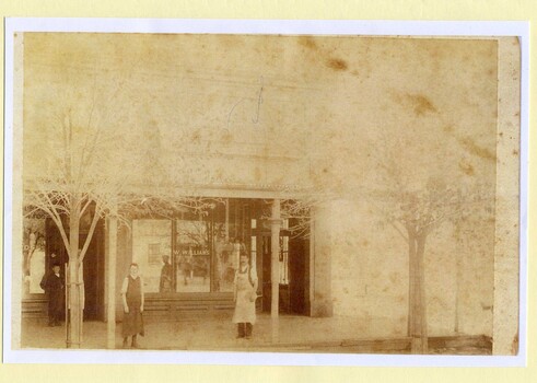 Four men in aprons standing under varanda in front of saddlery shop. Sign on window reads "W, Williams"