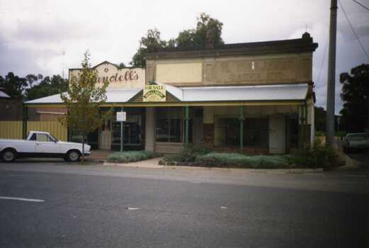 Two shop fronts. Left is Arundell's Shoe shop and right is William's Saddelery. Both brick building with large varanda in front.