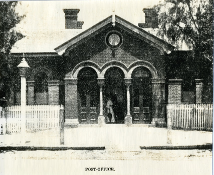 Brick building with three arched doorways. Woman in long frock carrying an umbrella standing in central doorway. White picket fence and lamp on footpath.
