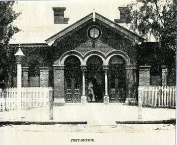Brick building with three arched doorways. Woman in long frock carrying an umbrella standing in central doorway. White picket fence and lamp on footpath.
