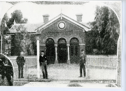 Three men standing in front of the brick postoffice. Has a white picket fence and three arches with columns.