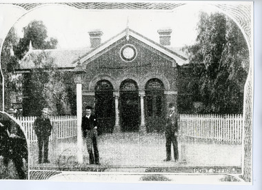 Three men standing in front of the brick postoffice. Has a white picket fence and three arches with columns.