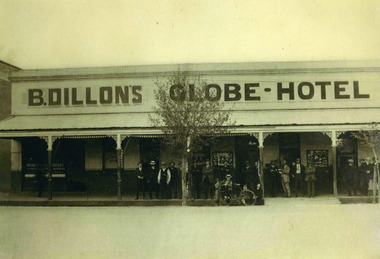 18 Adult males standing under varanda of B Dillon's Globe Hotel in suites and hats. Four boys in front of group, two sitting, two holding a bicycle.