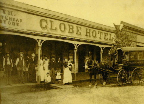 Large group of people standing outside the Globe hotel; four women, one holding a baby and four men. There is a little girl pushing a toy pram and a boy sitting. There is also a horse and buggy.