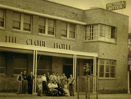 Group of 14 people standing and sitting on the verandah of the Globe Hotel. One man is playing a piano accordion and another has a small guitar. 