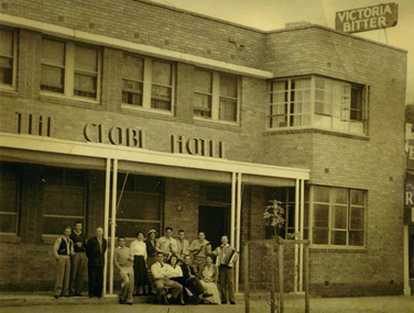 Group of 14 people standing and sitting on the verandah of the Globe Hotel. One man is playing a piano accordion and another has a small guitar. 