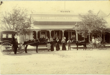 Klug's building. Four men standing on footpath, four women and a man holding a dog standing on the road. A horse and buggy either side of the group.