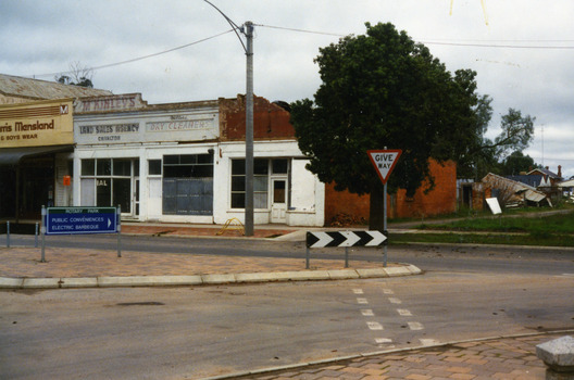 Shops at the west end of High St.Eddie Harris Mensland, Mickinley's Land Sales Agency, Hillier's Dr Cleaners and Manchester Unity. Sign on median strip pointing to Rotary Park.