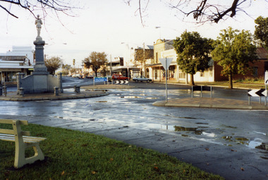 The road is wet. On the RH side , Eddie Harris Mensland, Paterson's FibreGlass factory, the Charlton Cinema, two story butcher shop.