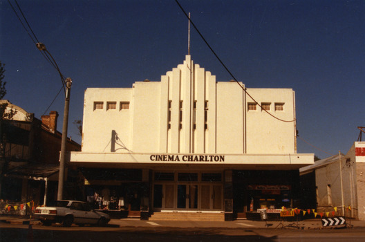 White building in an art deco style. White car parked in front. Dress shop and hairdressers . Roadwork happening.