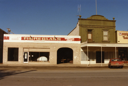 Green two storey building with verandah over the footpath and part of Mensland to the right. Red car parked in front of two storey building.