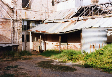 Rear of an old, brick two storey building. Building appears derelict and disused.Old rainwater tank on RH side, part of the roof is missing and the beams are exposed.
