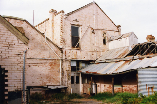 Rear of an old, brick two storey building. Building appears derelict and disused. Part of the roof is missing and the beams are exposed.