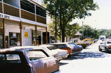 Two storey building with verandah over footpath and cast iron balcony on 2nd storey. Cars parked along the street. Large tree in centre of photo.